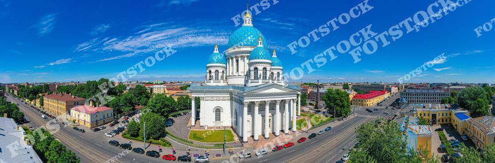 На фотографии изображён Троицкий собор — один из крупнейших православных храмов Санкт-Петербурга, выполненный в стиле ампир. Он выделяется своими ярко-голубыми куполами с золотыми звёздами. Собор расположен в Адмиралтейском районе и хорошо виден с высоты, окружённый старинной городской застройкой. Летнее солнце освещает белоснежный фасад и зелёные кроны деревьев. Снимок передаёт атмосферу ясного дня в центре Северной столицы.